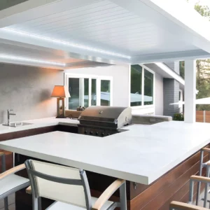 Modern outdoor kitchen sheltered under a durable Apollo louvered pergola, showcasing a stainless-steel grill and sleek countertops against a contemporary home backdrop.
