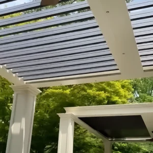 Close-up of a motorized louvered pergola with open bronze louvers, showcasing elegant sand-colored columns and a lush green background.