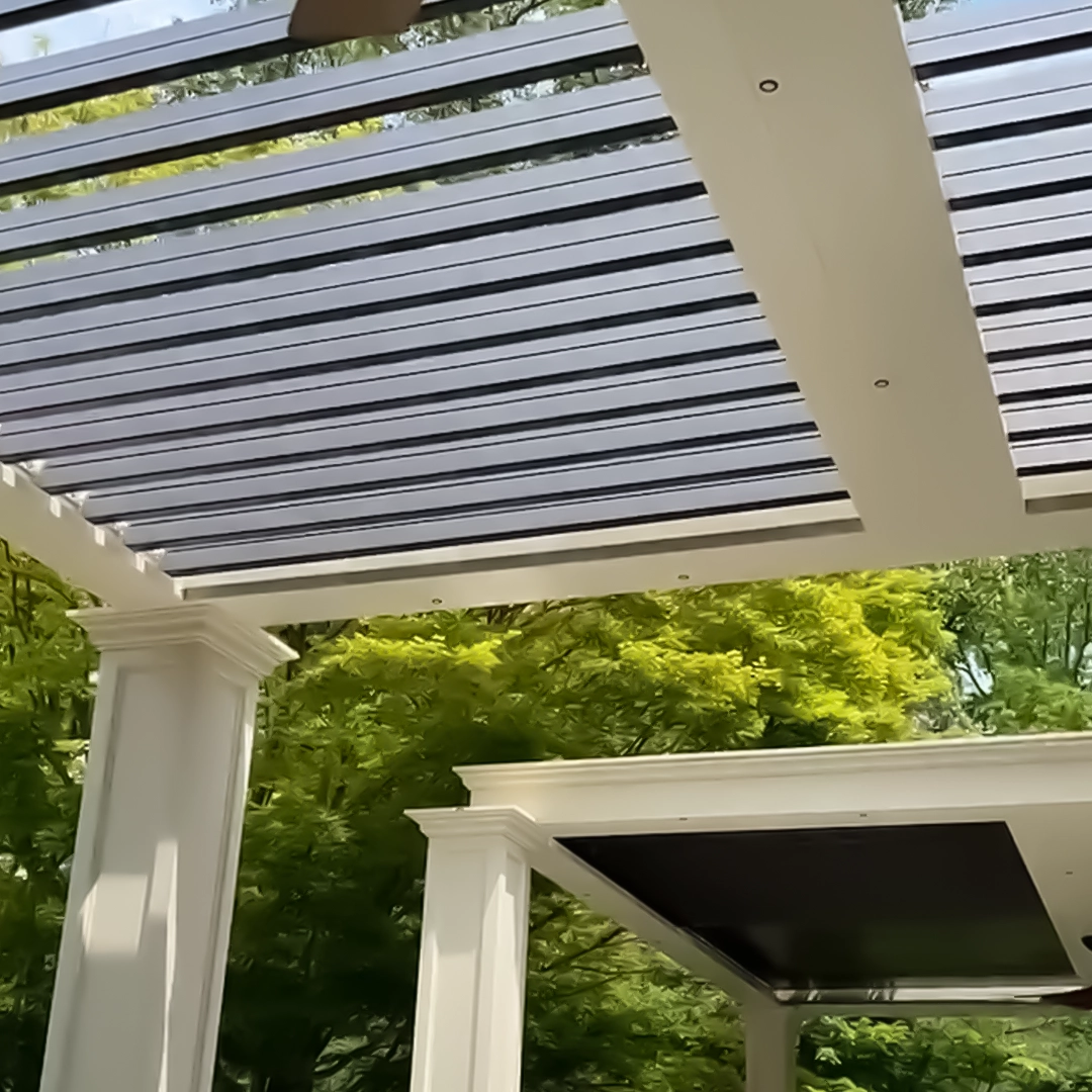 Close-up of a motorized louvered pergola with open bronze louvers, showcasing elegant sand-colored columns and a lush green background.