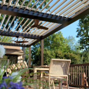 Outdoor dining area under a motorized louvered pergola with a ceiling fan, surrounded by lush greenery and a pool in the background.