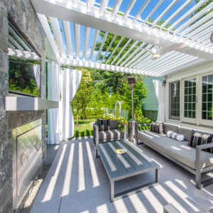 Outdoor entertainment area with a motorized louvered pergola, featuring a large stone fireplace, comfortable seating, and lush greenery.