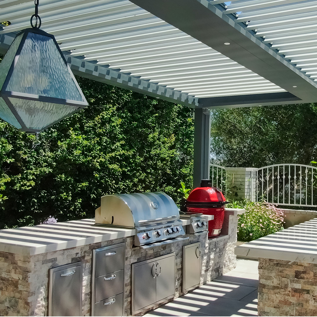 Outdoor kitchen with stainless steel appliances under a motorized louvered pergola, providing adjustable shade.