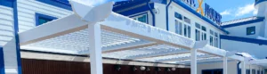 A snow-covered louvered pergola attached to a building with blue trim and windows, showing the structure's durability and ability to handle winter conditions.