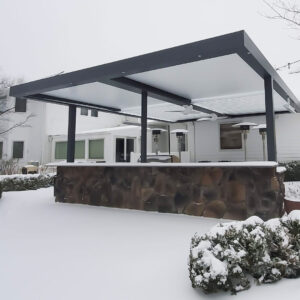 A modern louvered pergola covered in snow, featuring sleek black beams and posts, installed in a residential backyard with a built-in stone counter. The pergola is designed to withstand snow loads and harsh winter conditions.